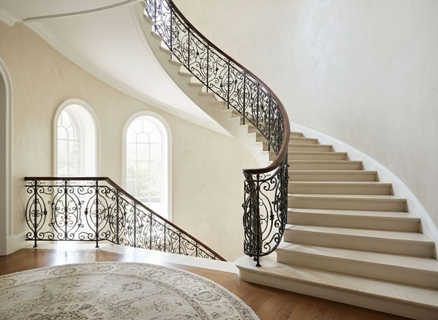 glaMUR Waende in W101 Elfenbein. Geschwungene Treppe mit kunstvollem Geländer, drei hohe Fenster im Hintergrund und ein runder Teppich auf dem Holzboden.