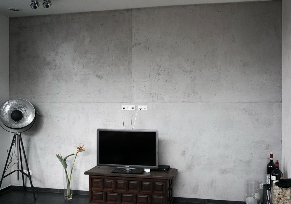 Wohnzimmer mit Betonwand, einem Fernseher auf einem Holztisch, einer Stehlampe links und einer Vase mit Blume.