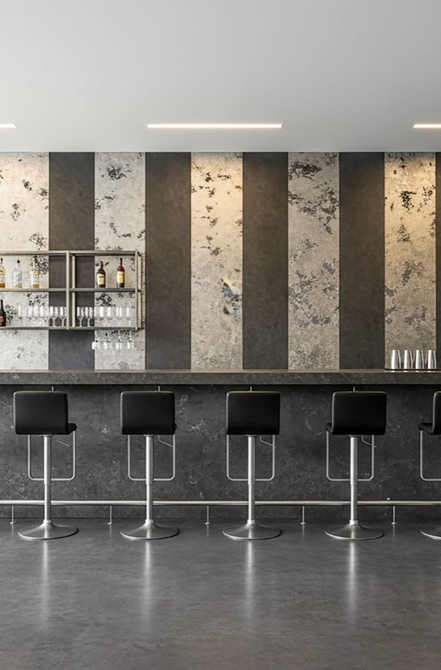 Eine moderne Bar mit acht schwarzen Barhockern vor einer Effektspachtel Wand mit abwechselnd Platin und Smoke vertikalen Streifen.